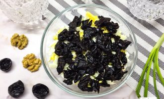 Нарезанный чернослив в миске, выложенный поверх яиц, на фоне кухонной салфетки, рядом ингредиенты салата — грецкие орехи, чернослив и зелёный лук.