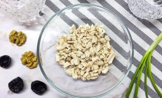 Отварное и нарезанное куриное филе в стеклянной миске, рядом грецкие орехи, чернослив и зеленый лук на фоне полосатой салфетки.
